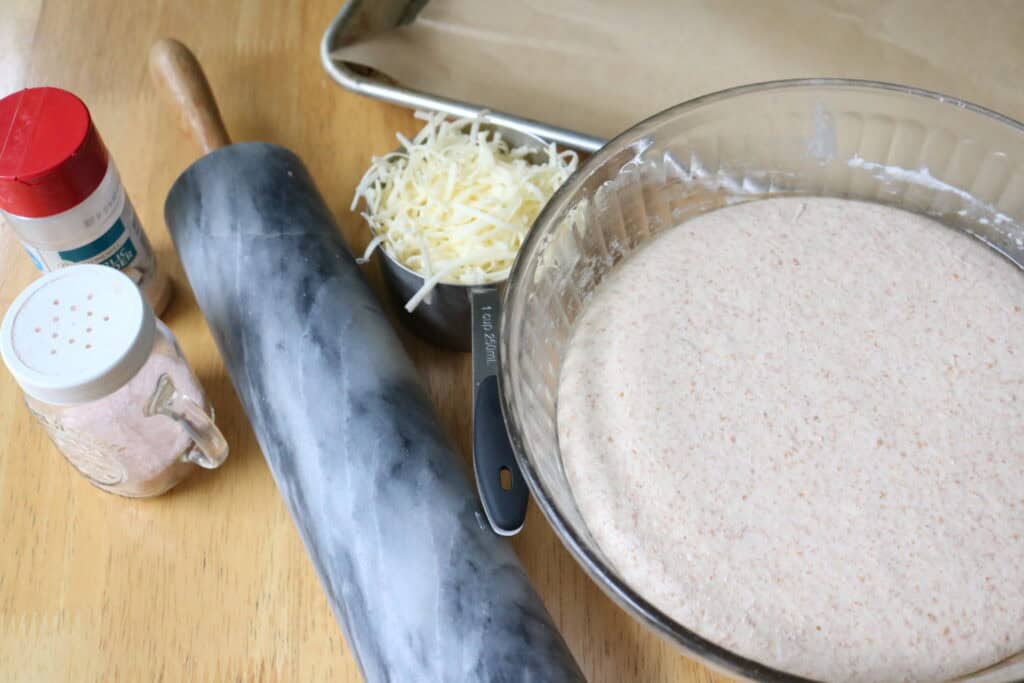 bowl of dough, baking sheet, rolling pin, grated cheese, and spice on a countertop
